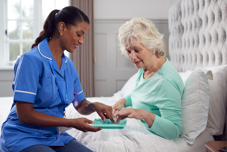 Female Care Worker In Uniform Helping Senior Woman At Home In Bed With Medication